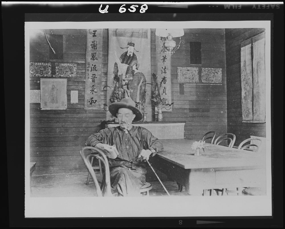 U658 [Historic photograph of a chinese man reading and smoking, in a decorated room]